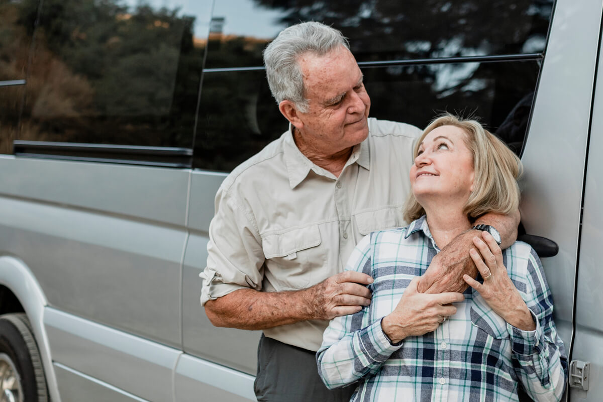 Un couple de retraités souriant devant leur camping-car profilé sur une aire de stationnement.